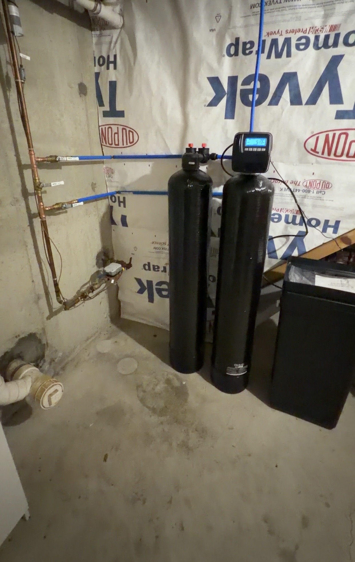 Two black treatment tanks connected with blue piping along a basement wall, with a brine tank at the side.