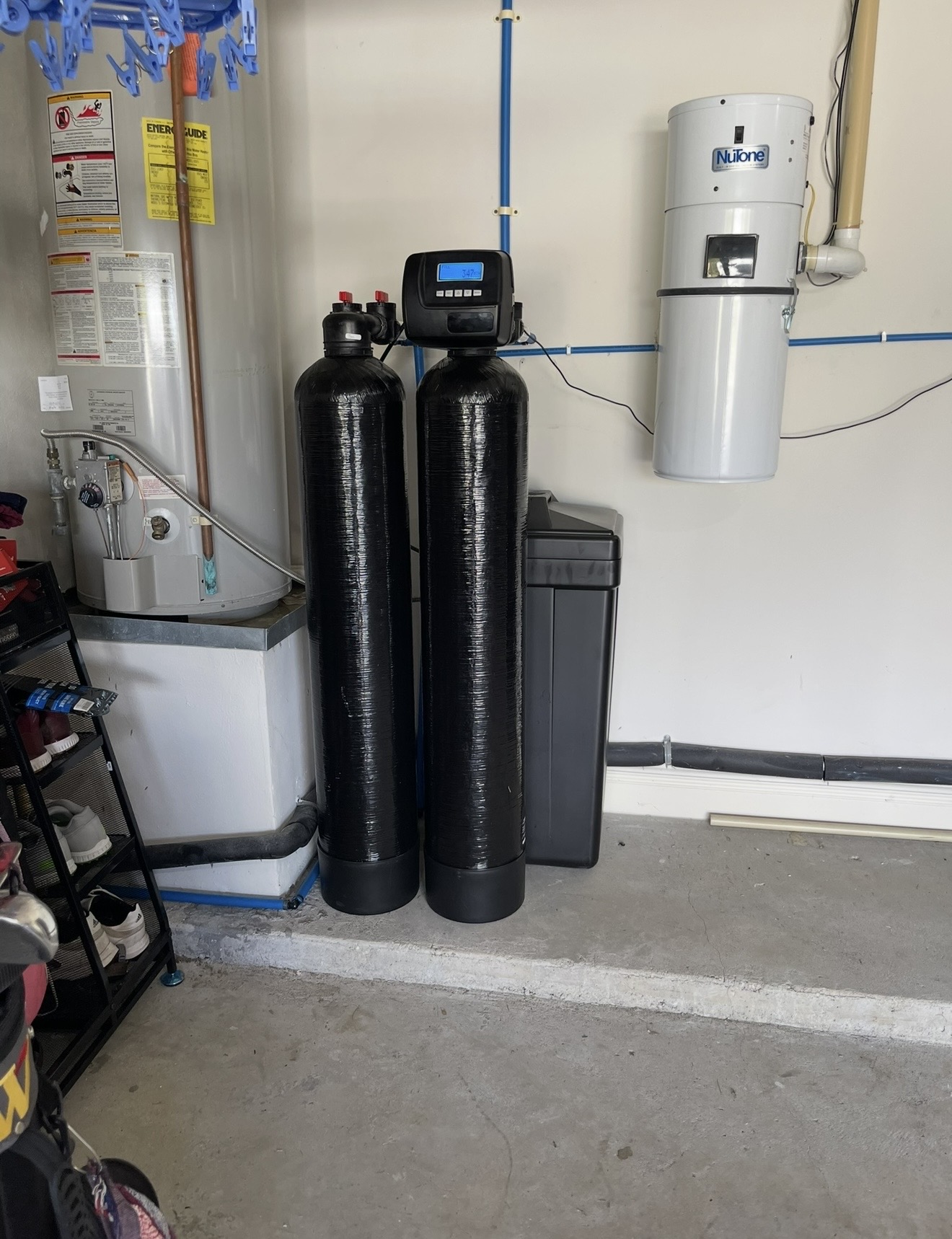 Two black water treatment tanks with a digital controller and a brine tank beside a garage water heater.